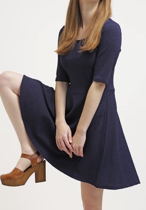 Femme portant une robe bleu marine texturée avec des manches au coude et des talons compensés marron, levant un genou avec les mains posées dessus.