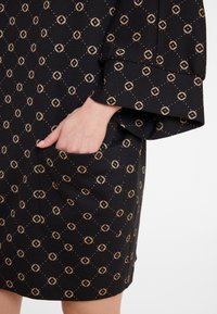 Zwarte jurk met een geometrisch patroon van beige diamanten en stippen, met een loose fit en een zijzak die in het ontwerp is geïntegreerd.