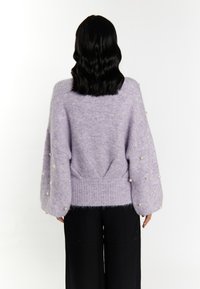 Pulover tricotat lila cu o textură pufos, mâneci supradimensionate împodobite cu perle albe și un tiv ribetat. Asortat cu pantaloni negri.