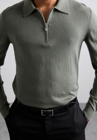 Maglione verde oliva con zip e polsini a coste, realizzato in morbido materiale a maglia. Abbinato a una cintura nera, presenta un aspetto pulito e moderno.