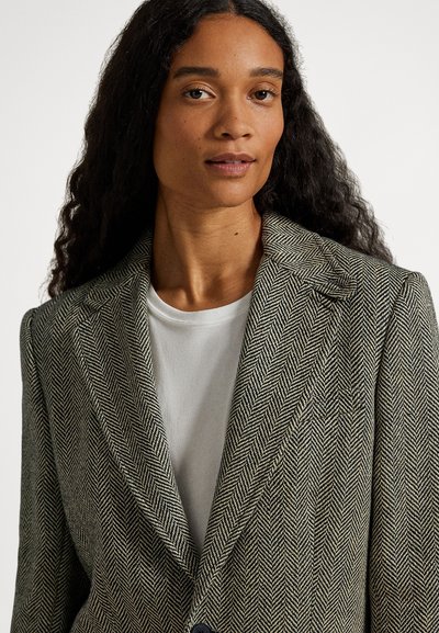 Kobieta z długimi kręconymi włosami, ubrana w marynarkę w jodełkę na białej koszulce z okrągłym dekoltem, zwrócona przodem z neutralnym wyrazem twarzy.