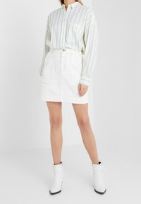 Witte button-up shirt met groene verticale strepen, gecombineerd met een witte denim rok. Model draagt witte enkellaarsjes met zwarte zolen.