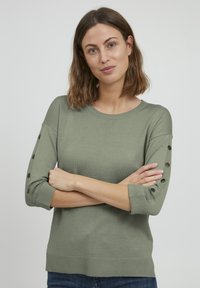 Pull à maille vert olive avec des manches trois-quarts, mettant en valeur des accents de boutons, un col rond et un ourlet droit. Texture douce et coupe décontractée.