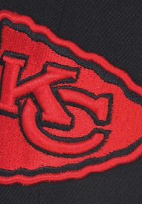 Rotes gesticktes Logo auf einer schwarzen Mütze, das ein stilisiertes "KC" innerhalb eines roten Rahmens zeigt, mit sichtbaren Texturen und Nähdetails.