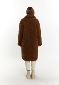 Brun fluffy oversized frakke med høj krave, lavet af tekstureret kunstpels. Båret over lyse bukser og ankelstøvler.