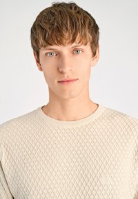 Beige värinen neulottu villapaita, jossa on teksturoitu timantinmuotoinen kuviointi, pyöreä pääntie ja resorit hihansuissa. Sileä kangas, jossa hienovaraisia yksityiskohtia.