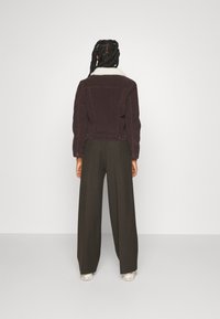 Una persona con capelli intrecciati si trova di fronte a una parete bianca indossando una giacca marrone scuro con colletto bianco, pantaloni larghi scuri e sneakers bianche.