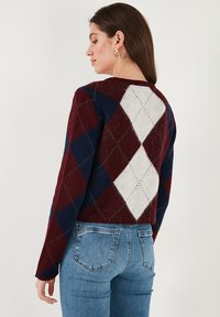 Knit sweater featuring a maroon, navy, and white diamond pattern, long sleeves, and a relaxed fit, paired with blue denim jeans.
