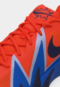 Φωτεινό πορτοκαλί αθλητικό παπούτσι με μπλε και λευκό σχεδιασμό με οδοντωτές γραμμές, μαύρο λογότυπο Nike και στυλιζαρισμένο σημάδι αστραπής στη γλώσσα.