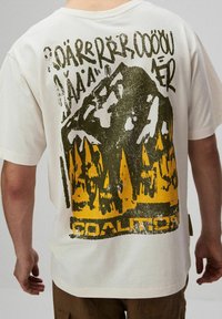 Biała koszulka z żółtą i czarną grafiką przedstawiającą góry oraz las, z postarzonym napisem powyżej i słowem "KOALICJA" poniżej z tyłu.