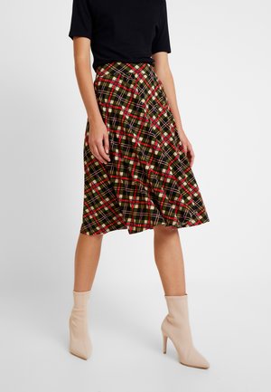 Femme portant une jupe écossaise aux couleurs vert, rouge et noir, longueur genou, un haut noir à manches courtes et des bottines beige à talons hauts.