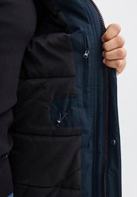 Marineblå isolert jakkeinteriør som viser en glidelåslomme og snor detalj. Stoffet har en glatt, matt tekstur.