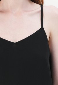 Haut noir sans manches avec un décolleté en V. Confectionné en tissu lisse. Fines bretelles menant à une coupe décontractée au niveau du corps. Légère brillance visible.