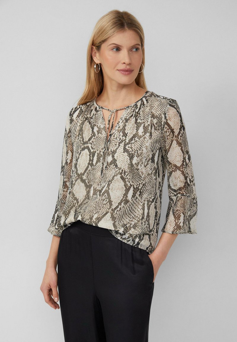 Blouse transparente avec un motif en peau de serpent dans des tons beige, noir et crème. Présente un décolleté en keyhole et des manches évasées trois-quarts.
