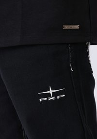 Zwart katoenen overhemd met een metalen label met de tekst "PROJECT X PAND." Zwarte broek met witte "PXP" borduursel en textuurstof.