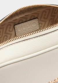 Valkoinen teksturoitu nahkalaukku kullanvärisellä vetoketjulla, jossa on vaaleanbeige sisävuori ja näkyvä brändilappu.