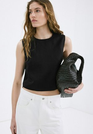 Femme aux cheveux ondulés portant un haut noir sans manches et un pantalon blanc, tenant un grand sac à main noir tressé sur un fond blanc uni.