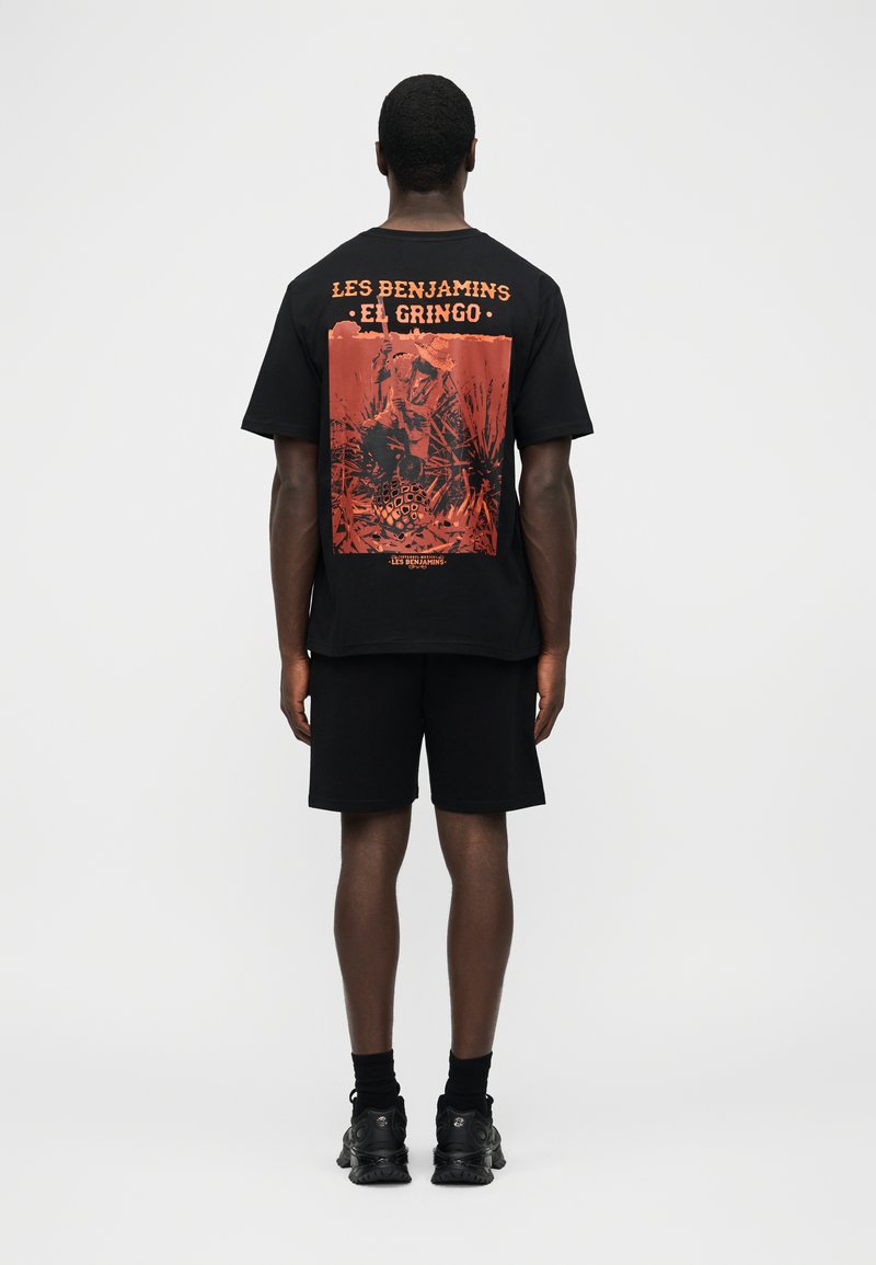 Άνδρας που φοράει μαύρη μπλούζα και σορτς, η πίσω πλευρά της μπλούζας δείχνει πορτοκαλί γραφικό και κείμενο "Les Benjamins El Gringo" σε απλό φόντο.