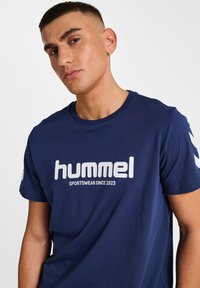 Navyblaue Baumwoll-T-Shirt mit einem großen weißen "hummel"-Logo und dem Text "SPORTSWEAR SINCE 1923". Kurze Ärmel mit kontrastierenden Streifen.