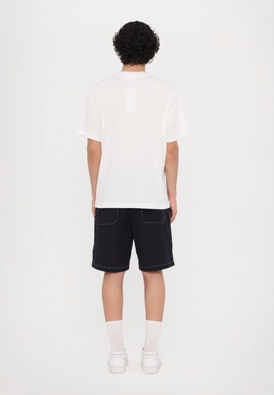 Tricou alb din bumbac cu mâneci scurte, asociat cu pantaloni scurți negri cu cusături albe. Vederea prezintă partea din spate a ținutei.