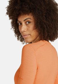 Oranje langekleurige top met een ronde hals en een gestructureerde stof. Heeft stikseldetails en een klein logo op de linkermouw.