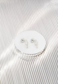 Silvriga blixtformade studörhängen som visas på en cirkulär reflekterande yta. Slät finish med subtil textur. Minimalistisk design.