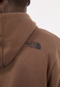 Personne portant un sweat à capuche marron avec le logo The North Face imprimé en noir sur le haut du dos, près de la capuche.