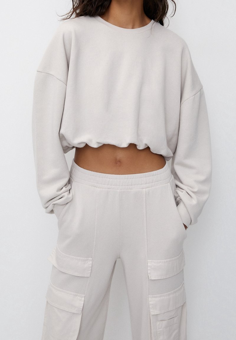 Femme portant un sweat-shirt court beige clair et un pantalon cargo assorti, les mains dans les poches, sur un fond uni.