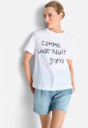 Vrouw met kort blond haar draagt een wit T-shirt met Franse tekst "Comme une nuit d'été" en lichtblauwe spijkershorts, staand tegen een witte muur.