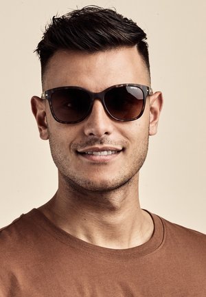 Lunettes de soleil avec des verres foncés et une monture écaille de tortue. Forme carrée, rehaussée par un subtil accent en métal sur les tempes.