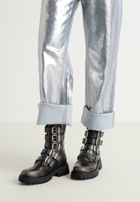 Bocanci negri din piele pentru gleznă, cu mai multe curele argintii cu catarame, cu tălpi texturate groase, asortați cu pantaloni argintii lucioși, cu manșete.