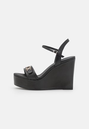Sort kile-sandal med ankelrem, åben tå, platformsål og detaljer med metalspænde på frontremmen.