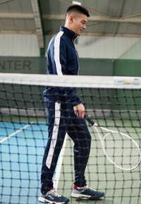 Marineblauer Trainingsanzug mit weißen Streifen, bestehend aus einer Jacke mit Reißverschluss und passend geschnittenen Hosen. Hält einen Tennisschläger auf einem Platz. Strukturiertes Oberflächen sichtbar.