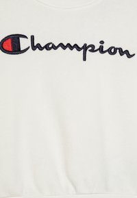 Sudadera Champion en tejido crema con logo bordado en azul marino y rojo. Cuello redondo clásico y ribete acanalado para mayor estructura.