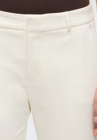 Nahaufnahme von cremefarbenen Hosen mit vorderen Taschen und Gürtelschlaufen, getragen mit einem dunkelbraunen Oberteil, wobei ein Teil einer Hand an der Seite zu sehen ist.