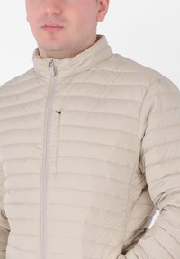 Giacca puffer beige con motivo a trapuntatura orizzontale, colletto alto e zip frontale completa. Presenta un piccolo logo sulla manica.