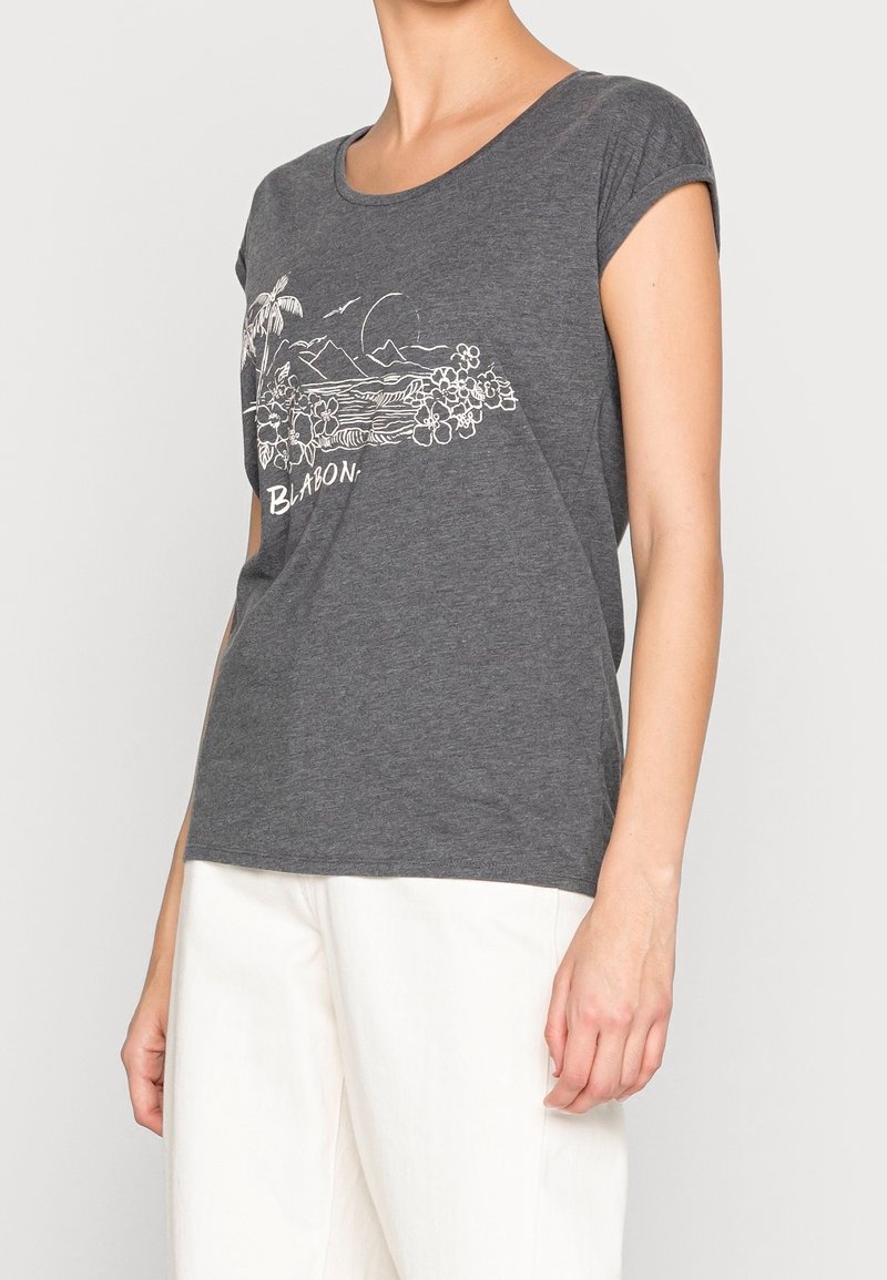Personne portant un t-shirt gris foncé avec un motif blanc d'île tropicale et le mot « BLABON », associé à un pantalon blanc.
