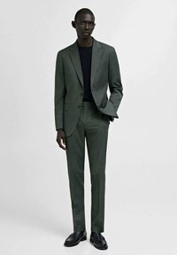 Grön skräddarsydd kostym med en enfaldig kavaj, smal passform på byxorna och en enkel svart skjorta. Avslutas med svarta loafers. Slät textur.