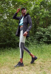 Chaqueta ligera negra, camiseta azul verdosa, leggings grises con acentos negros y zapatillas deportivas negras. Modelo caminando en un entorno boscosa verde.