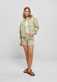 Chaqueta de mezclilla oversize beige con dos bolsillos en el pecho, camiseta blanca y pantalones cortos beige claros, combinados con sandalias planas de mimbre marrones.