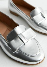 Srebrne metaliczne loafersy z kokardką z przodu, okrągłym noskiem oraz niskim brązowym obcasem. Gładka tekstura i elegancki design.