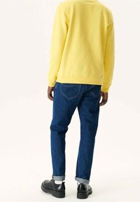 Personne portant un haut jaune à manches longues, un jean bleu retroussé, des chaussettes blanches et des chaussures noires, debout et tournée de dos sur un fond uni.