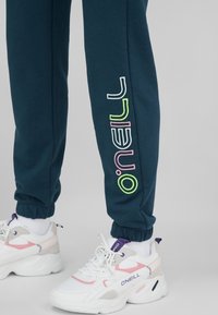 Tyrkysové tepláky s elastickými manžetami mají na noze barevné logo "O'Neill". Kombinují se s robustními bílými teniskami s růžovými detaily.