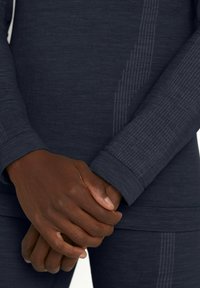 Ciemnoszara bluzka z długim rękawem wykonana z teksturowanej tkaniny. Posiada prążkowane mankiety i pionowe linie dla dodatkowego efektu. Ręce złożone na wysokości talii.