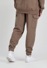 Smeđe trenirke s bočnim cargo džepovima, elastičnim manžetama i opuštenim krojem, uparene s bijelim Nike tenisicama. Glatka tekstura tkanine.