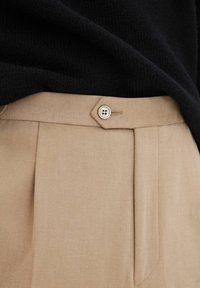 Beige skräddarsydda byxor med framsydda veck, silverfärgad knappstängning och slät tygkvalitet. Bärs med en svart stickad topp.