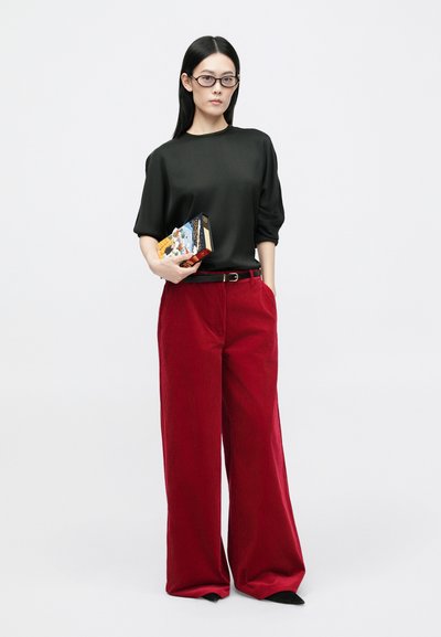 Zwarte top met korte mouwen, rode corduroy wijde broek, zwarte riem, houdend een doos met kleurrijk ontwerp, gedragen met zwarte puntige schoenen.