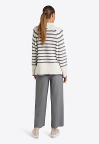 Gestreifter Pullover in Creme und Grau, mit geripptem Saum, kombiniert mit grauen, weit geschnittenen, plissierten Hosen. Das Modell zeigt die Rückansicht, die ein Knopfdetail offenbart.