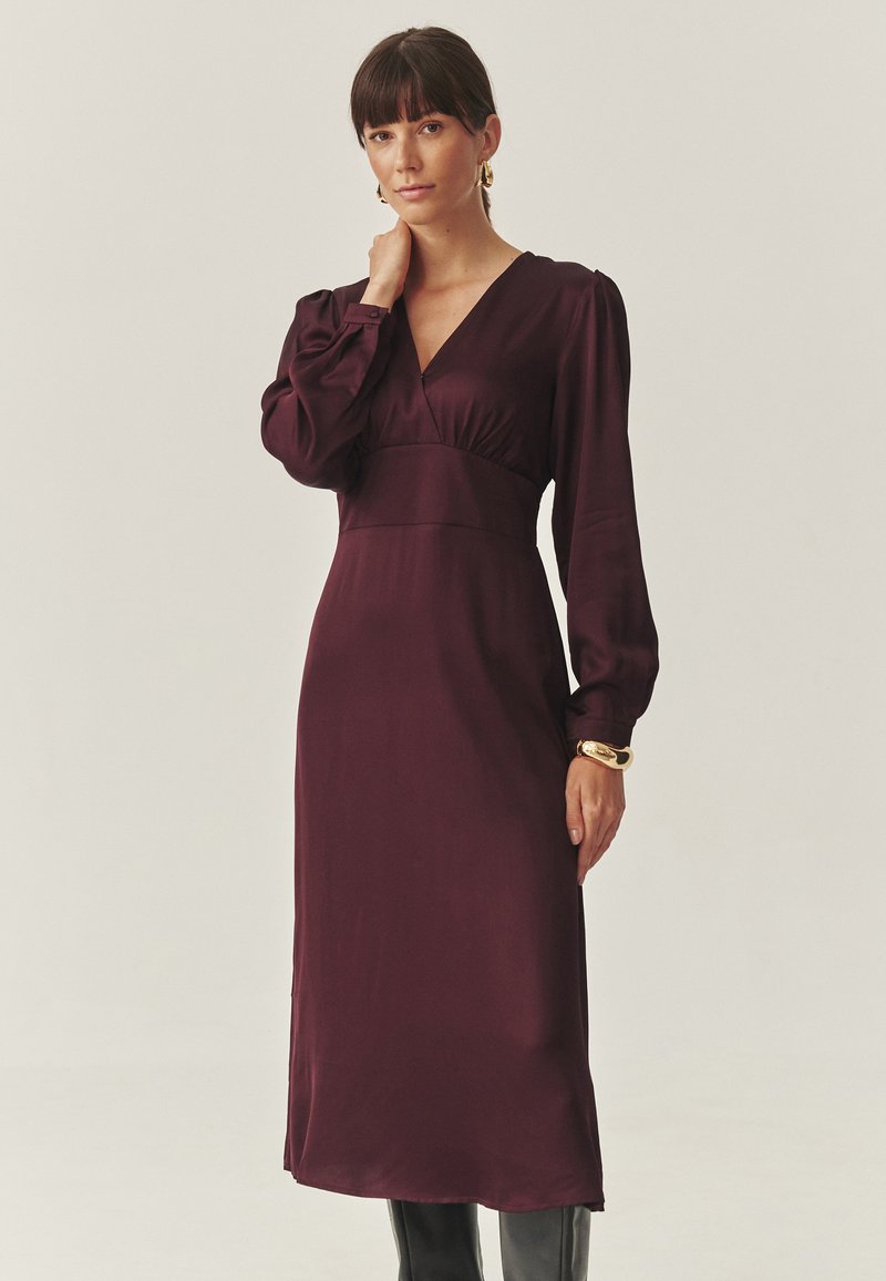 Rochie din satin prune cu decolteu adânc în V, mâneci lungi tip puff și talie strânsă. Designul pune în evidență o siluetă fluidă și o textură netedă.