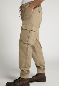 Pantaloni cargo beige in cotone, con tasche laterali, vita elasticizzata con cordino e polsini aderenti. Abbinati a stivali marroni con lacci.